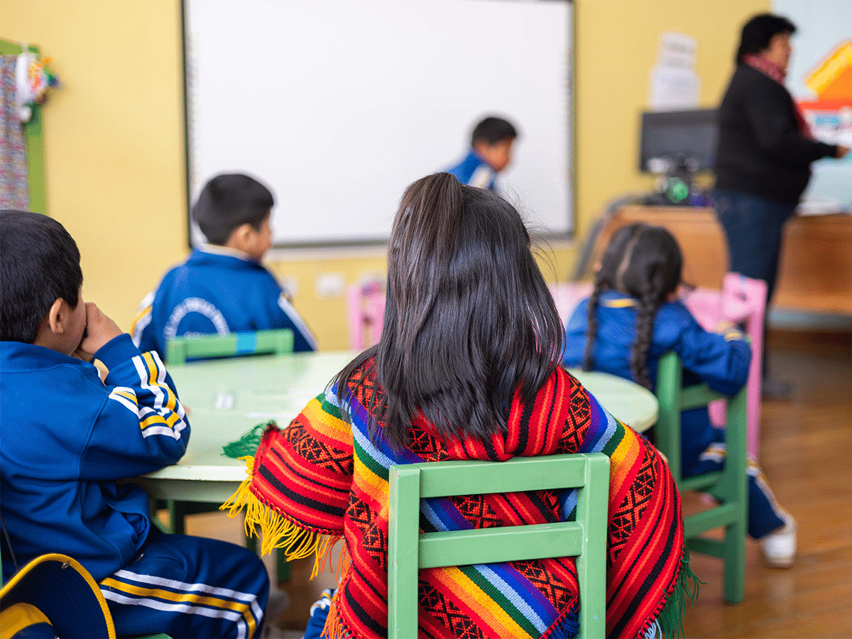 An over-the-shoulder view of young students sitting at tables in a classroom in Peru, facing forward toward a teacher. A girl in the foreground wears a vibrant, rainbow-striped traditional Andean poncho over her shoulders, while the other children wear blue and yellow school tracksuits.