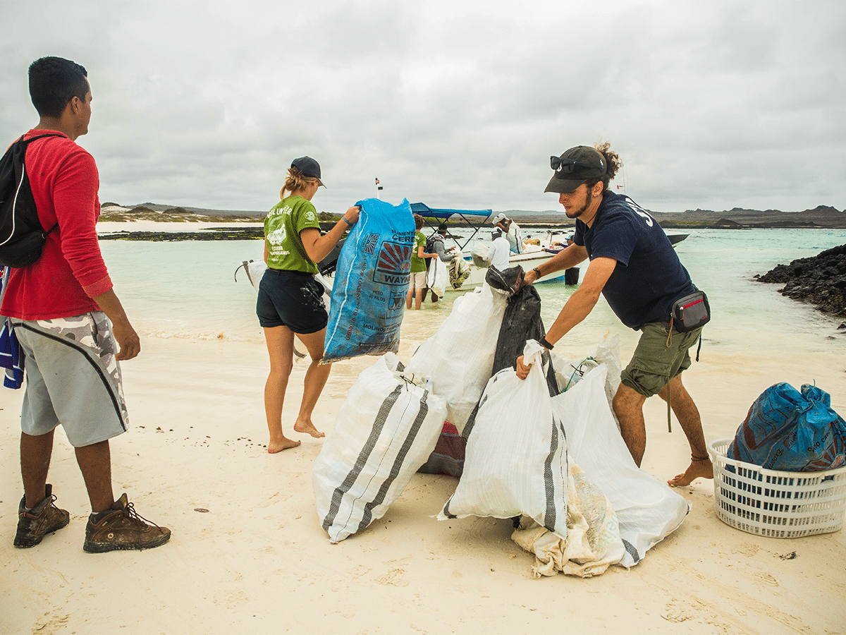 Volunteers participate in a beach cleanup on a sandy shore in the Galapagos. A man in a dark t-shirt lifts large, filled woven sacks, while a woman in a green shirt carries a blue plastic bag toward a small boat waiting in the turquoise water under an overcast sky.