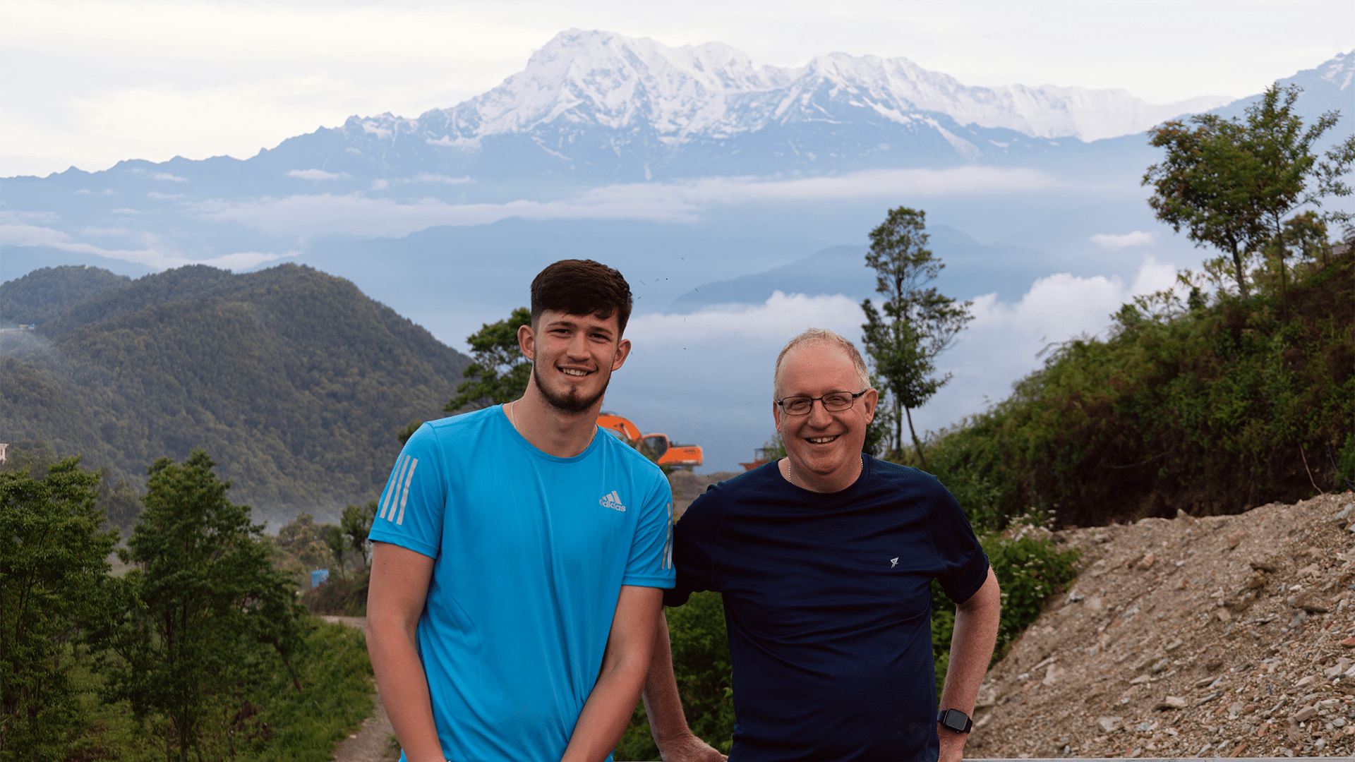 Two smiling men stand side-by-side in front of a metal railing, posing against a panoramic mountain backdrop. The younger man on the left wears a bright blue athletic t-shirt, while the older man on the right wears a dark navy blue t-shirt and glasses. In the background, green hills roll down into a layer of low-lying white clouds, above which looms a massive, snow-capped mountain range under a pale sky.