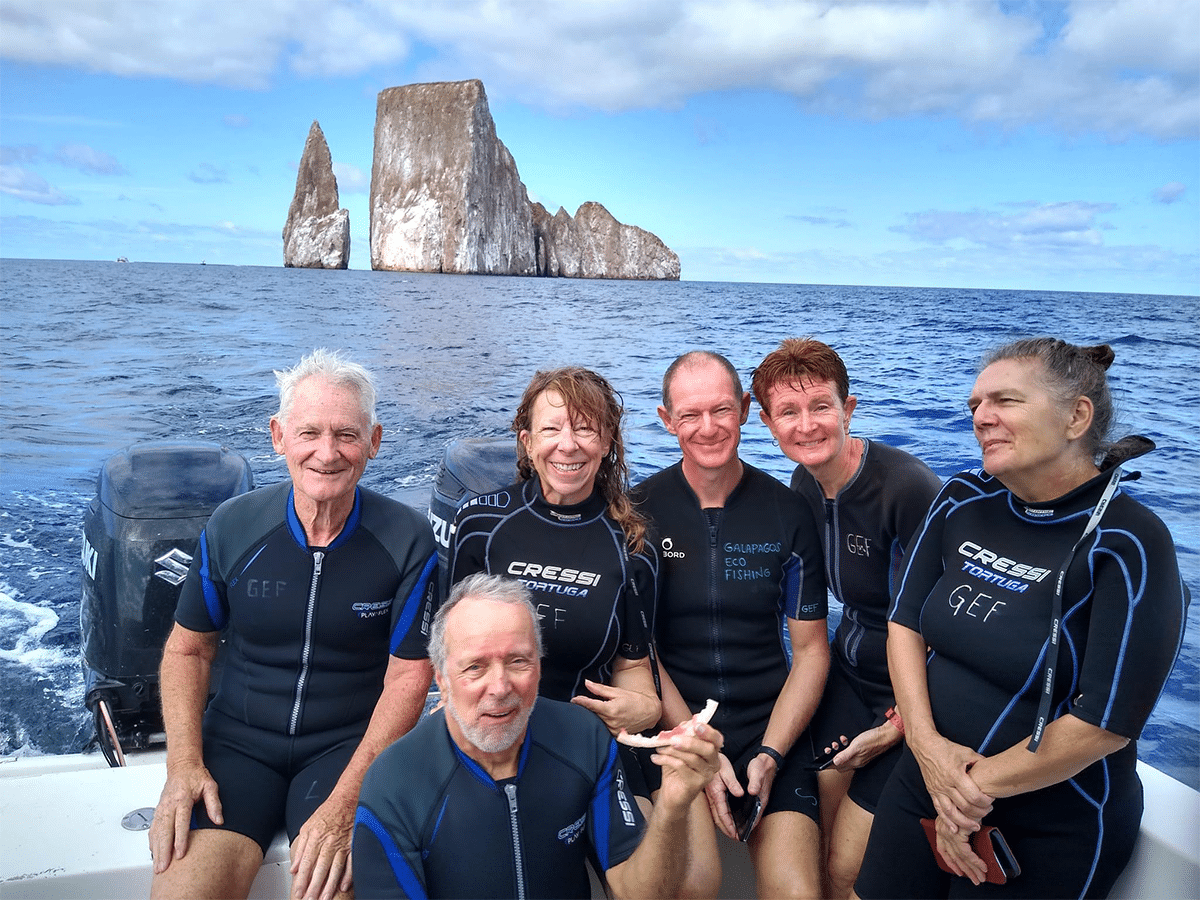 A group of six people wearing black wetsuits smiles while sitting on a boat on the ocean, with the prominent rock formations of Kicker Rock (León Dormido) visible in the background under a partly cloudy sky in the Galápagos Islands.