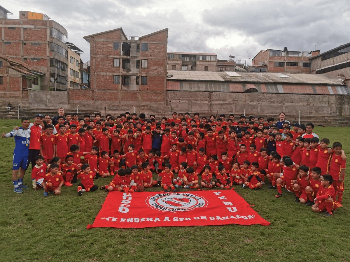 A large group of children and coaches pose together on a green soccer field in Peru. The children are wearing matching red and yellow uniforms, while a large red banner with a white circular logo and the text "TE ENSEÑA A SER UN GANADOR" is laid on the grass in front of them, with urban brick buildings under a cloudy sky in the background.