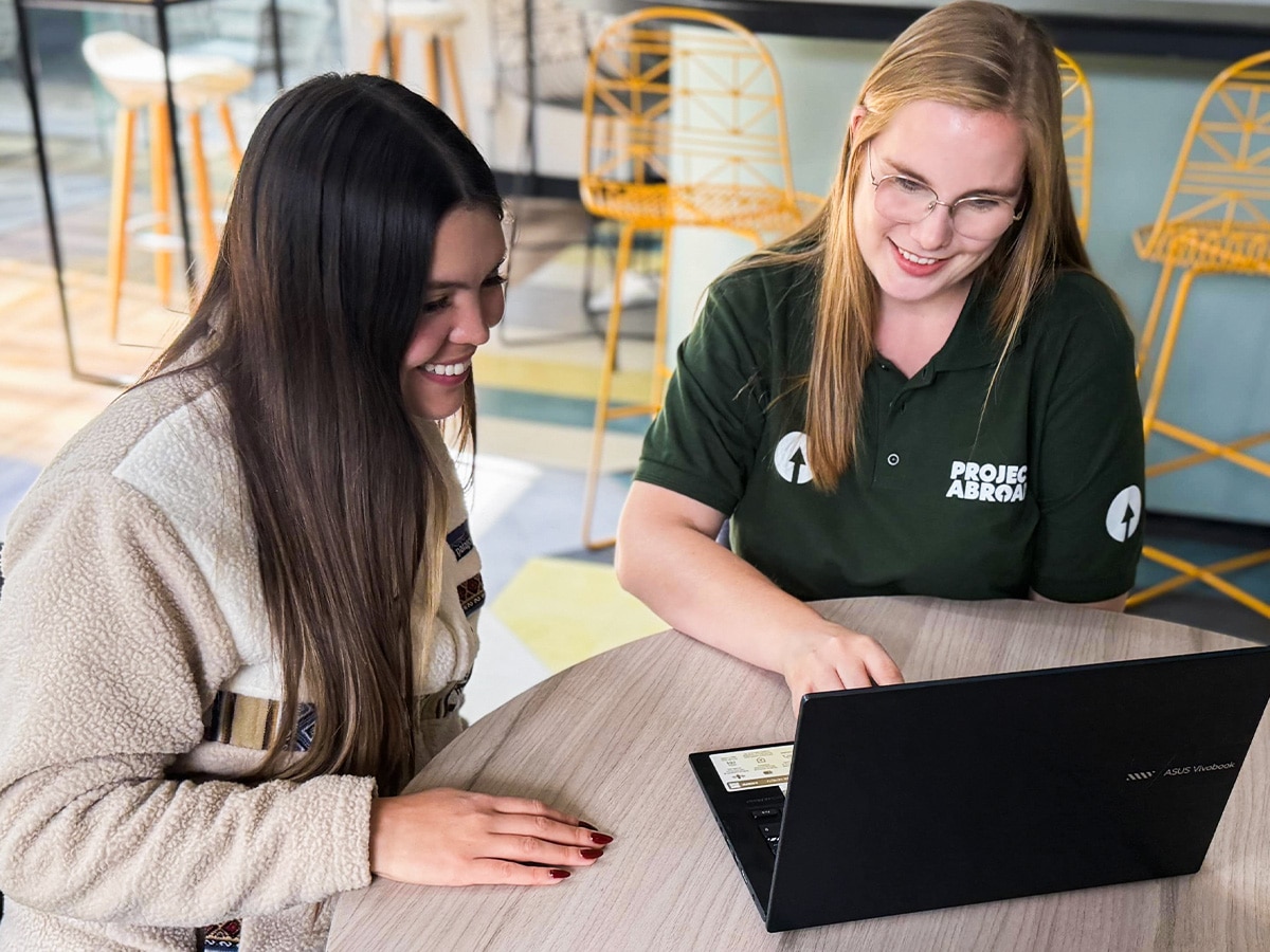 A Projects Abroad staff member sitting at a wooden table with a volunteer, both smiling as they look at a laptop screen together.