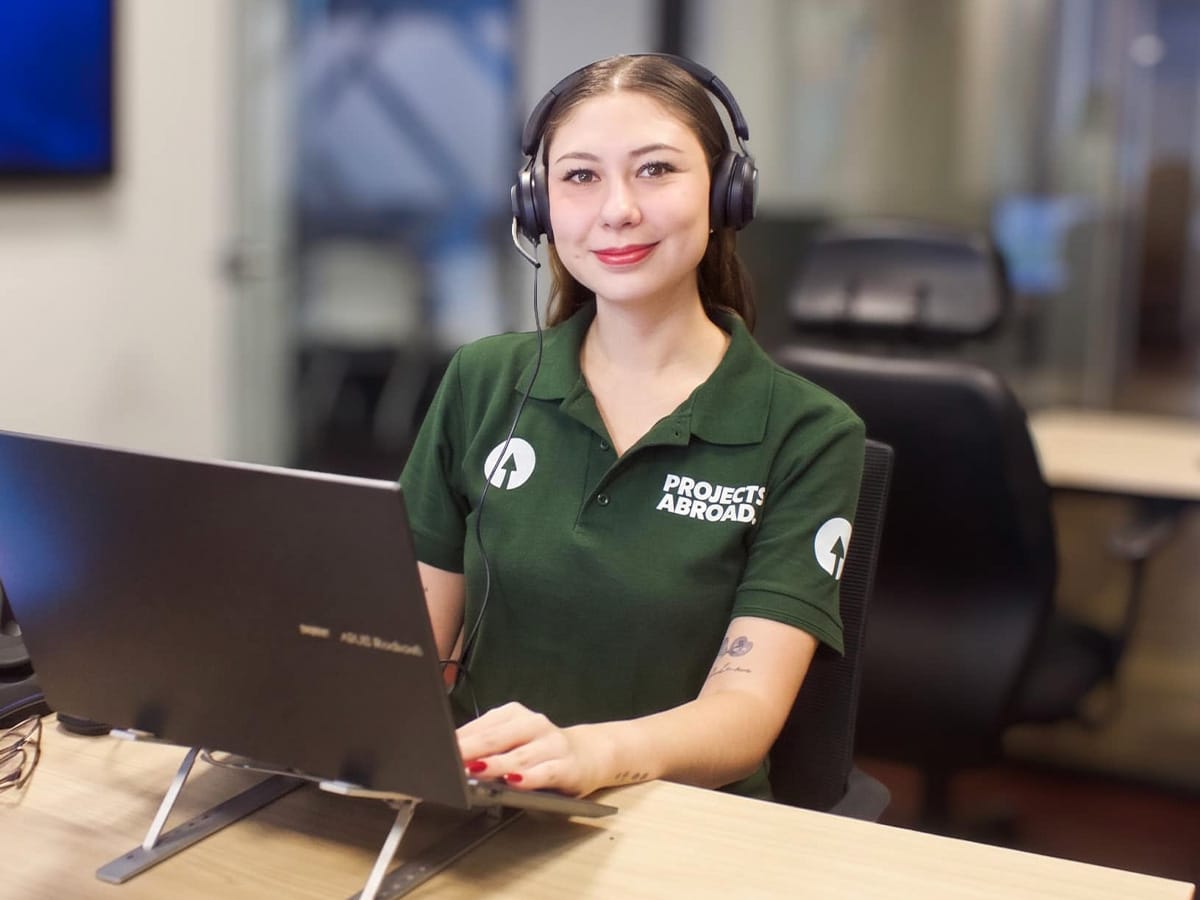 A smiling Projects Abroad staff member wearing a headset and dark green polo shirt, sitting at a desk with a laptop.